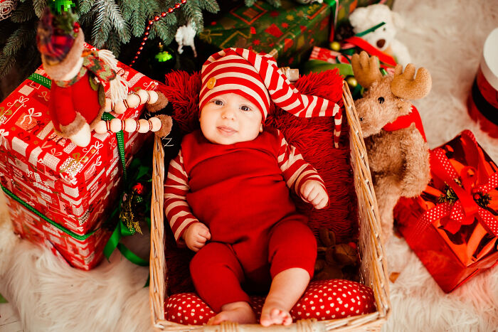 Baby dressed in red lying in basket under Christmas tree surrounded by holiday gifts and decorations.