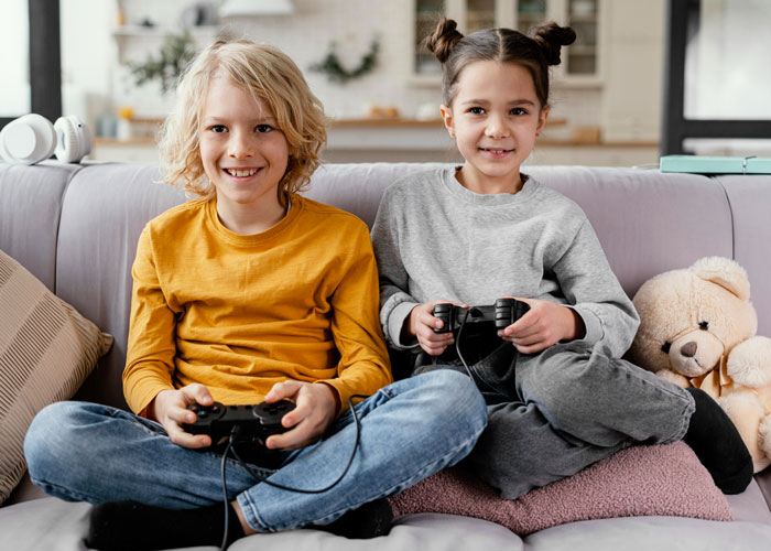 Two kids playing video games on couch, illustrating family babysitting expectations and demands for payment.