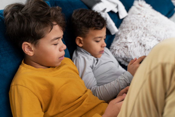 Two kids relaxing on a couch, focused on their devices while babysit sister watches during hockey day.