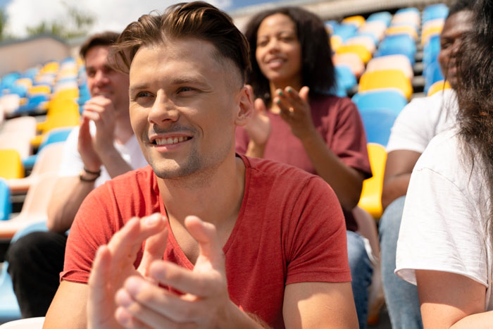 Young man babysitting sister's kids watching a hockey game, smiling and clapping in outdoor stadium seating.