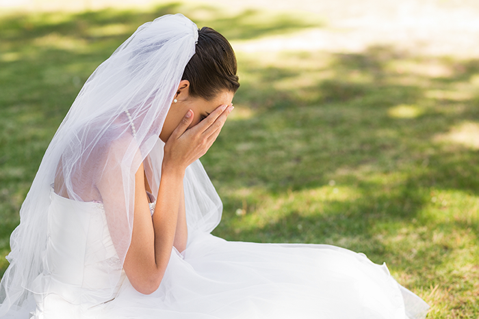 Bride in wedding dress sitting outdoors covering her face, upset after discovering groom cheated with her mom.