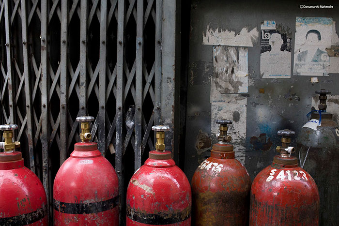 Row of rusty red gas cylinders lined up against a metal gate and dirty wall, illustrating chaotic good themes in urban settings.