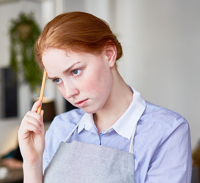 Young woman wearing apron holding pencil to head, appearing thoughtful, representing chaotic good moments shared by people.