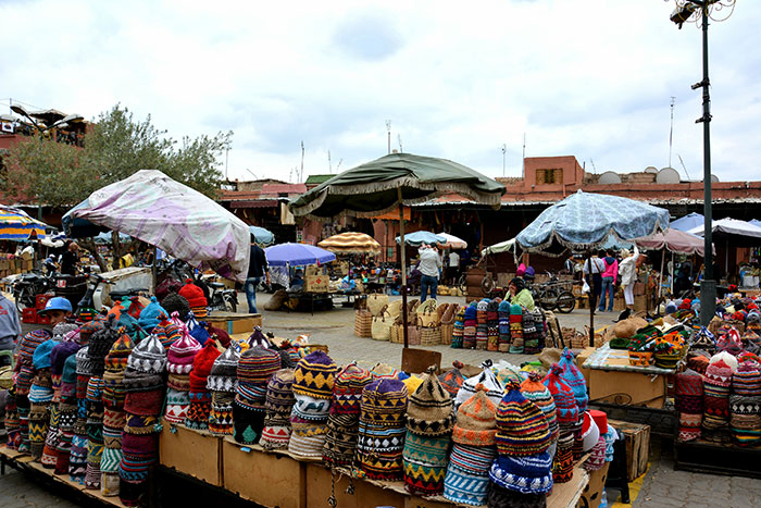 Colorful hats displayed at an outdoor market with people and umbrellas, capturing chaotic good moments in daily life.