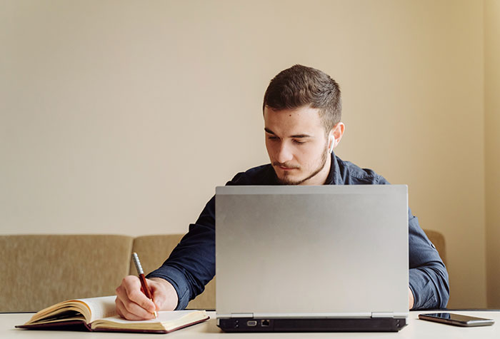 Young man working on chaotic good content creation, writing notes while using a laptop in a cozy indoor setting.