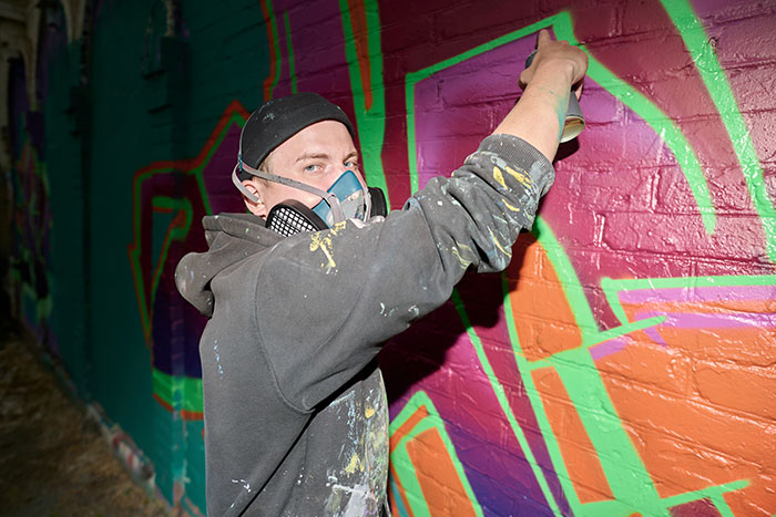 Artist wearing a mask spray painting colorful chaotic good graffiti on a brick wall at night in an urban environment.