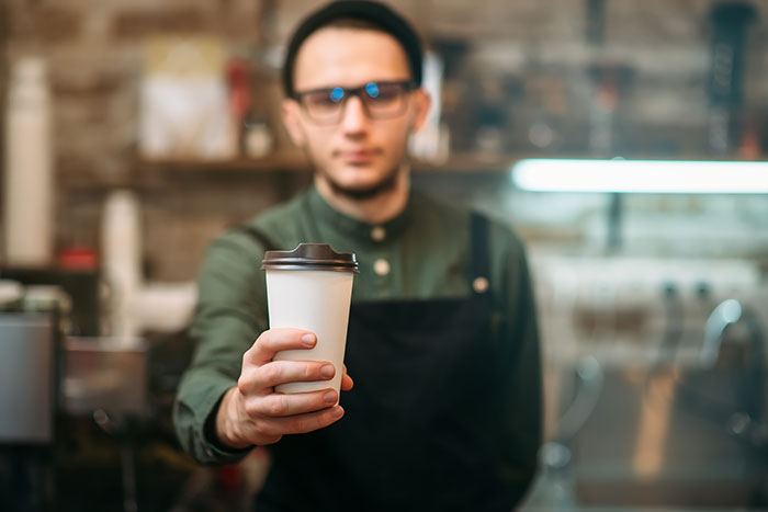 Barista in black apron offering a coffee cup in a cozy coffee shop, representing chaotic good moments.