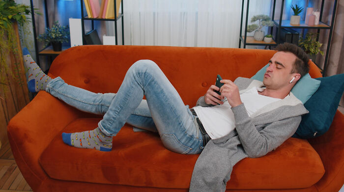 Young man relaxing on an orange sofa, using smartphone and wearing earbuds, illustrating biggest bullet dodged concept.