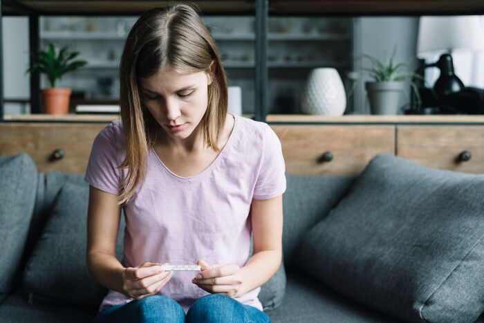Young woman sitting on a couch, looking at a thermometer, representing the biggest bullet dodged without realizing it.