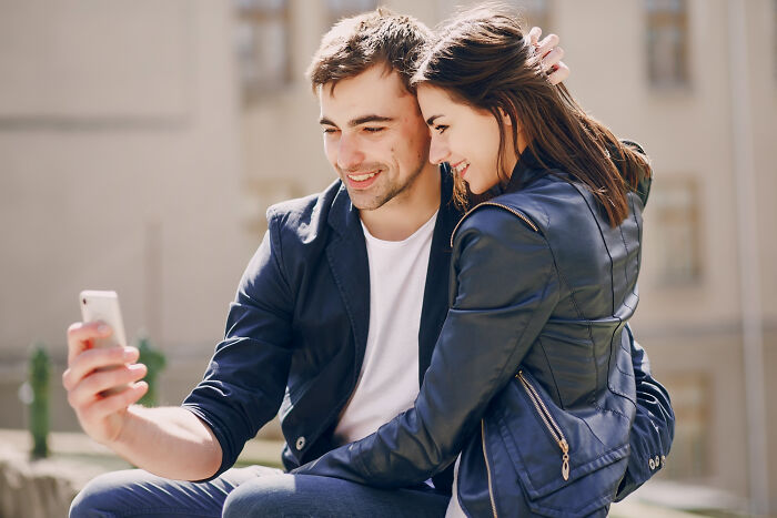 A happy young couple sitting outdoors, smiling and taking a selfie, representing biggest bullet dodged moments.