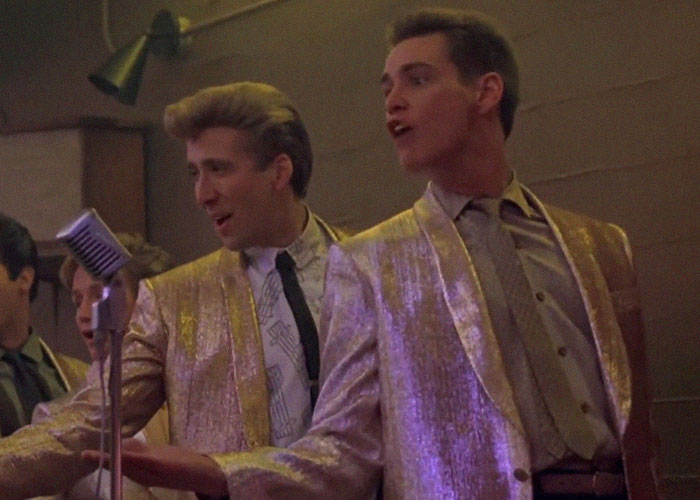 Actores en escena con trajes brillantes interpretando momentos de traición y backstabbing en Hollywood durante una presentación.