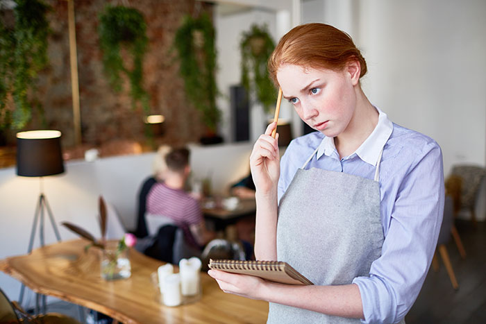 Young waitress wearing apron thoughtfully holding pencil and notepad in restaurant with customers dining in background Young waitress wearing apron thoughtfully holding pencil and notepad in restaurant with customers dining in background