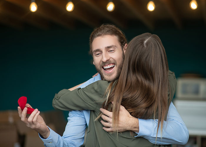 Man holding open ring box hugging woman, capturing moment of engagement during a joyful celebration.