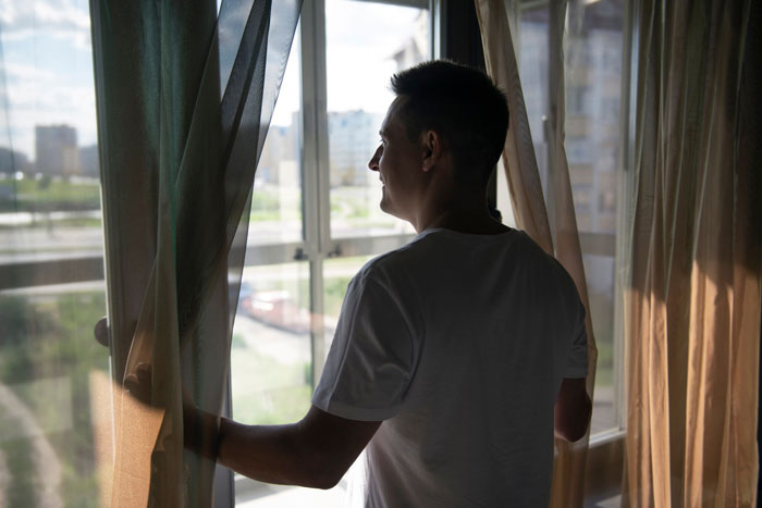 Man in white t-shirt standing by window with curtains, watching outside in a residential area near fire hydrant and driveway.