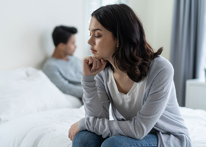 Woman looks upset sitting on bed while man in background appears distant, highlighting future child health concerns after a lie exposed. Woman looks upset sitting on bed while man in background appears distant, highlighting future child health concerns after a lie exposed.