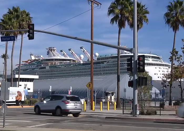 Large cruise ship docked at harbor with palm trees and moving cars at intersection under traffic lights.