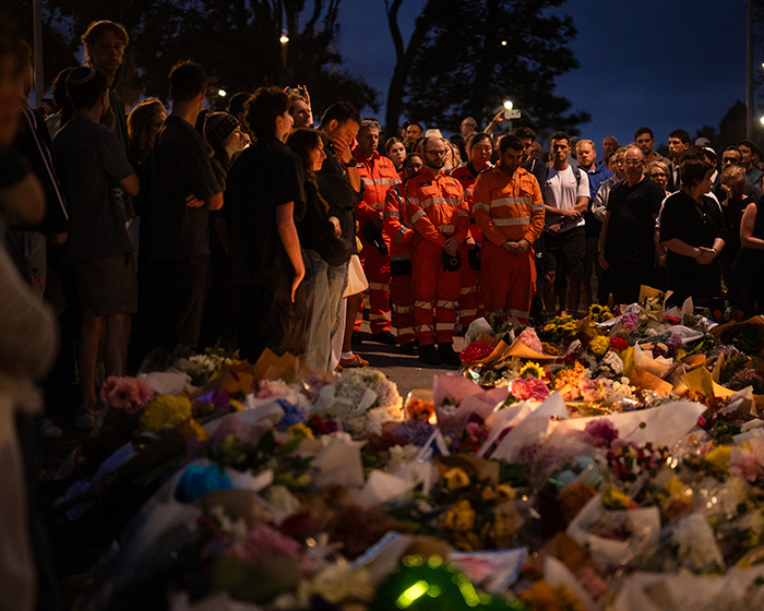 Crowd gathers at a memorial on Bondi Beach, honoring hero who confronted attacker, with emergency responders present at night.