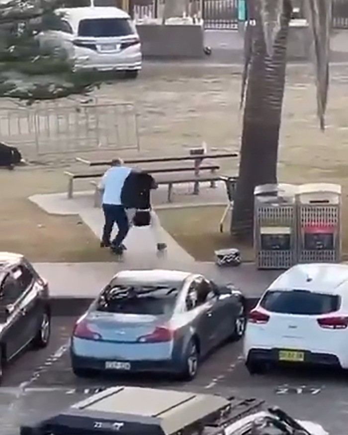 Man confronting attacker near skate park at Bondi Beach, with parked cars and picnic bench in background.