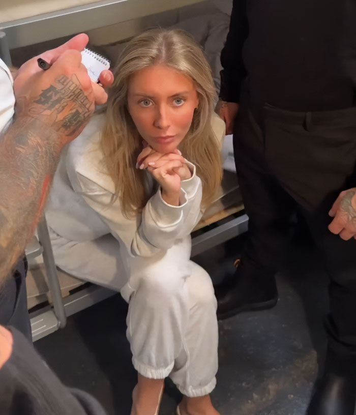Bonnie Blue sitting in a white outfit, looking defiant while surrounded by tattooed men in a dimly lit room.