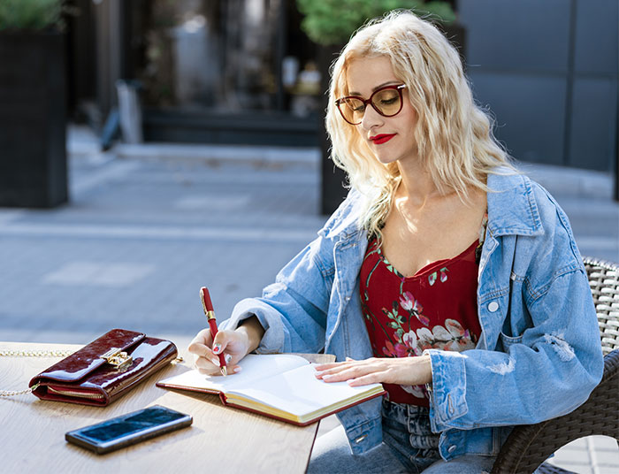 Woman writing novel outdoors, wearing glasses and denim jacket, seeking advice after boyfriend calls the police over her writing.