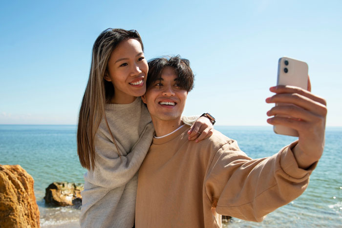 Young couple taking a selfie at the beach, boyfriend editing face every photo with a smile and affectionate pose.