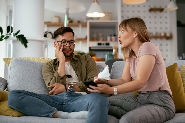A frustrated boyfriend editing face on every photo while sitting on a couch with a woman talking to him at home.