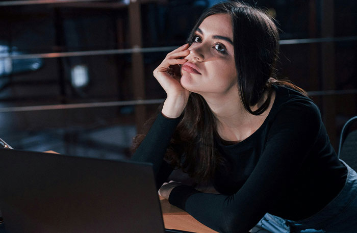 Young woman looking thoughtful while sitting at a desk with a laptop, reflecting on playing video games to unwind.