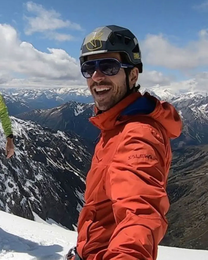 Man in orange jacket and helmet taking selfie on Austrian mountain, linked to boyfriend who abandoned woman story.