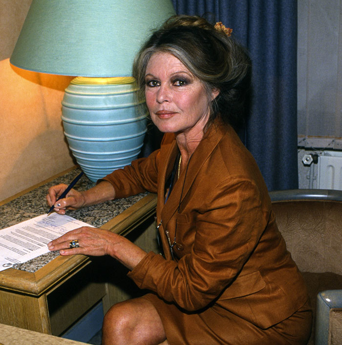 Brigitte Bardot sitting at desk signing papers with a lamp behind her, related to backlash over Chappell Roan post. Brigitte Bardot sitting at desk signing papers with a lamp behind her, related to backlash over Chappell Roan post.