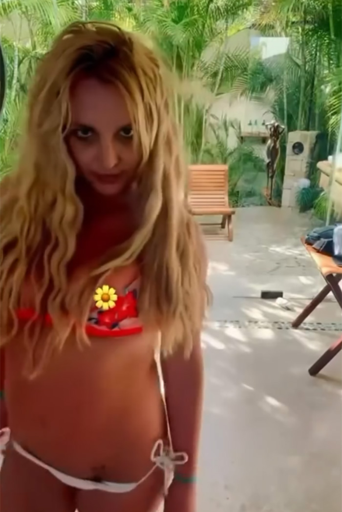 Britney Spears in a red bikini with a slip, standing outdoors near tropical plants and wooden furniture.
