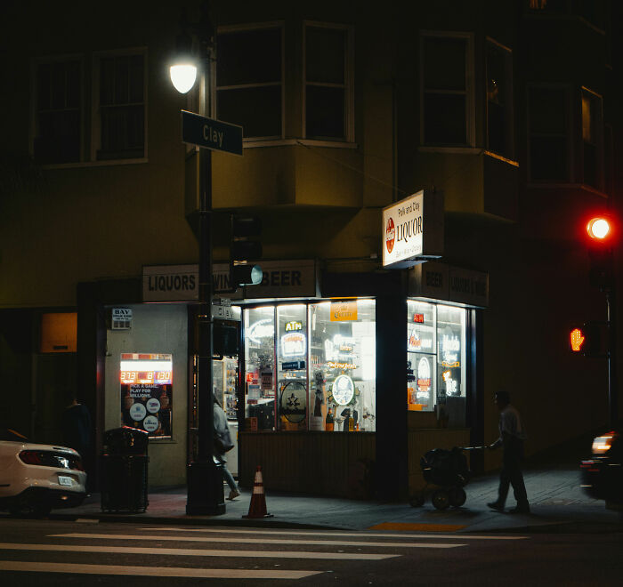 Nighttime view of a liquor store on a city corner with illuminated signs and pedestrians nearby related to raccoon raid.