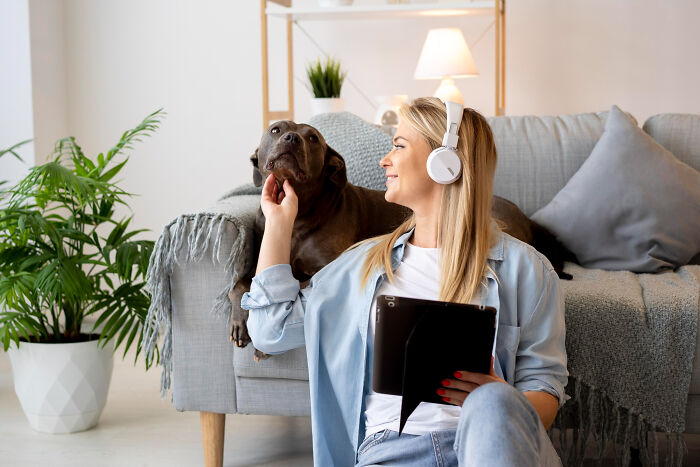 Woman wearing headphones, holding a tablet, smiling at a dog on the couch in a cozy living room setting.