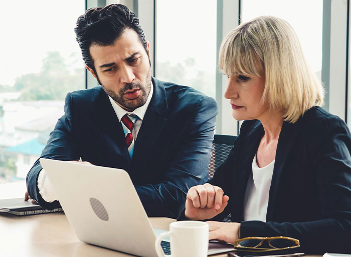 Man talking to woman looking weirded out in office setting with laptop and coffee cup on table, work mom coworker tension
