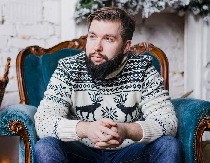 Bearded guy in a patterned sweater sitting thoughtfully in a vintage chair after siblings share surprise plans.