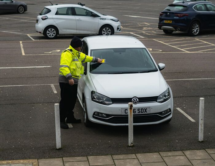 Parking enforcement officer placing a ticket on a white car in a nearly empty parking lot, showing signs of entitlement.