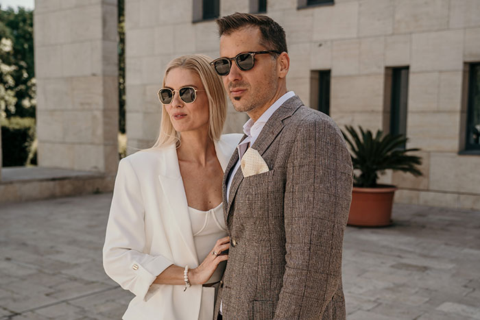 Stylish wealthy couple wearing sunglasses and elegant outfits outside a modern building symbolizing rich people lifestyle.