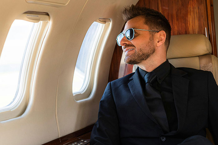 Man wearing sunglasses and a black suit sitting inside a private jet, illustrating rich people lifestyle realities.