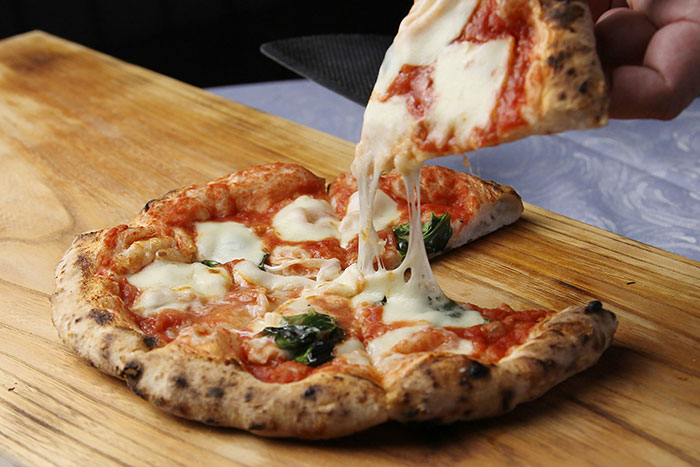 A slice of cheese pizza being lifted from a wood-fired pizza on a wooden board, showcasing melted cheese pull.