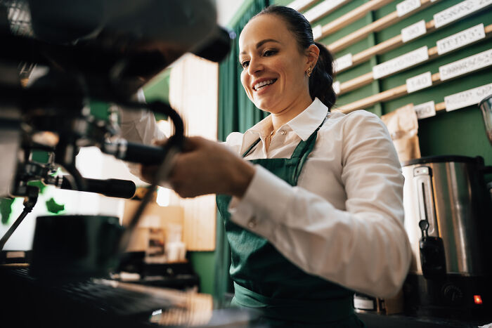 Smiling barista in a green apron making coffee in a cafe, representing people who became rich out of nowhere.