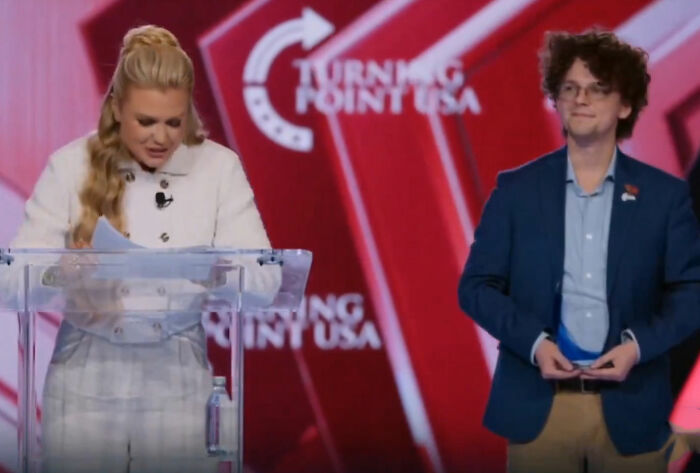 Erika Kirk making an awkward Freudian slip at a Turning Point USA event, standing beside a young man on stage.