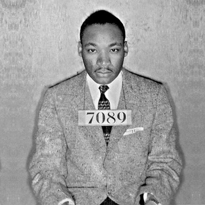 Black and white mugshot of a man wearing a suit and tie, holding an arrest number, representing celeb mugshot taken after arrest.