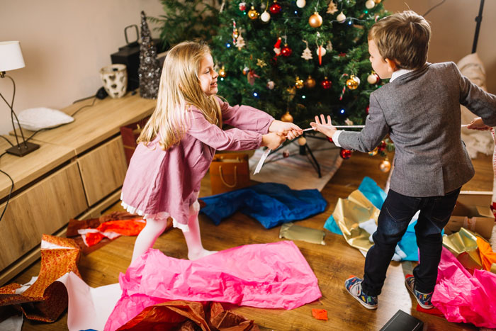 Two unruly children tugging a ribbon near a decorated Christmas tree with torn wrapping paper on the floor.