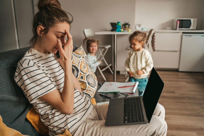 Stressed aunt ignoring unruly niblings playing in background with laptop on her lap in a cozy living room.