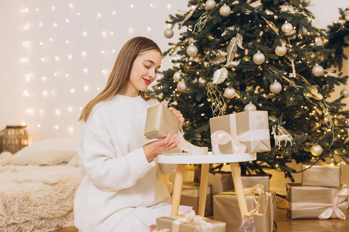 Woman in white sweater wrapping Christmas gifts by decorated tree, illustrating aunt no Christmas gifts for unruly niblings.