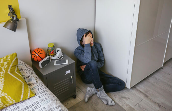 Sad child sitting on the floor in a hoodie with hand on face, near bed and nightstand, depicting a Christmas nanny kid bite moment.