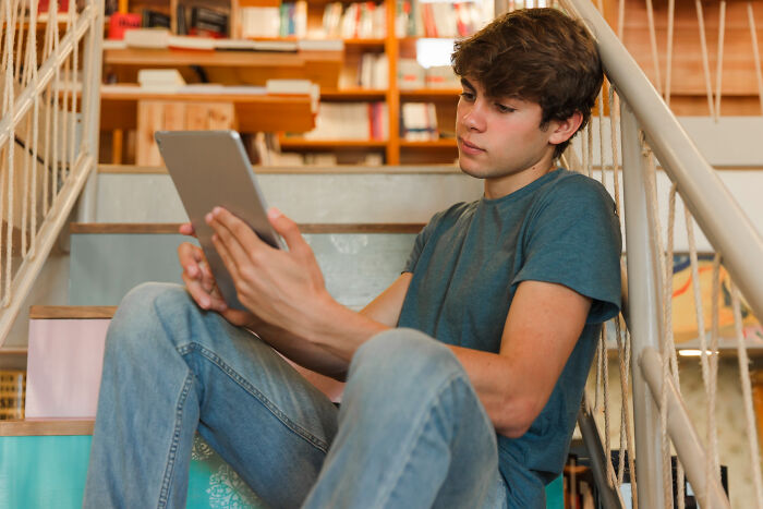 Teen boy sitting on stairs reading a tablet, looking thoughtful with bookshelves behind, related to Christmas nanny kid bite.