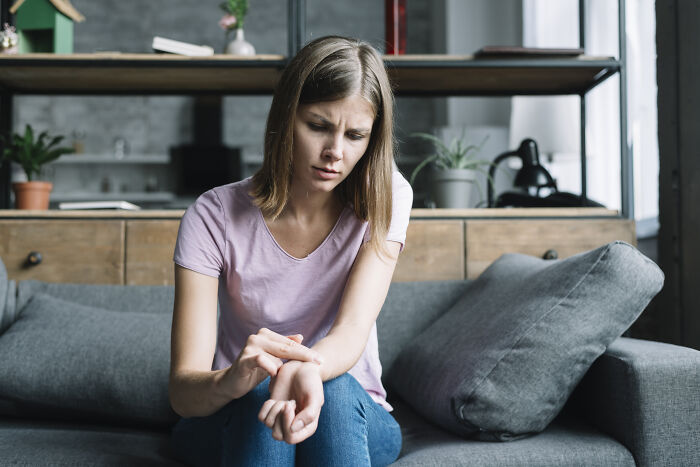 Worried nanny examining red bite marks on wrist while sitting on couch in a modern living room setting.