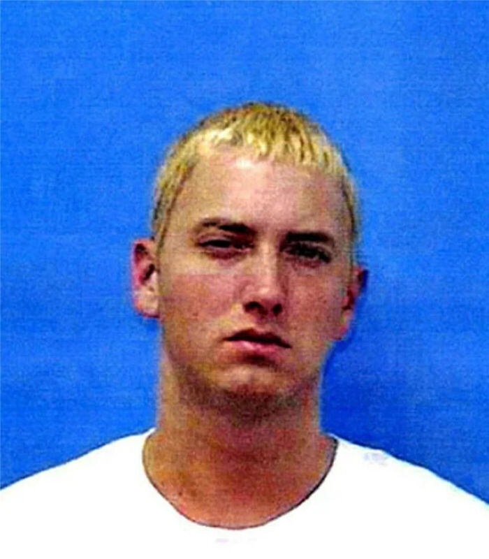 Young man with short blonde hair and white shirt posing for a celebrity mugshot after being arrested on blue background