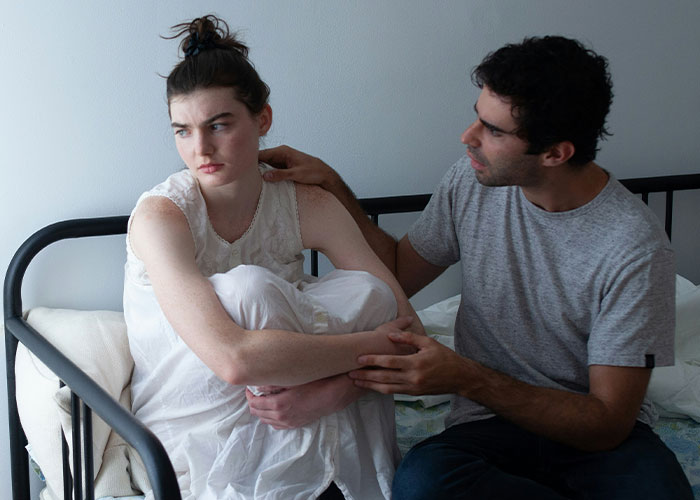 Couple in tense conversation on bed, woman upset and avoiding eye contact, man trying to comfort her after argument.