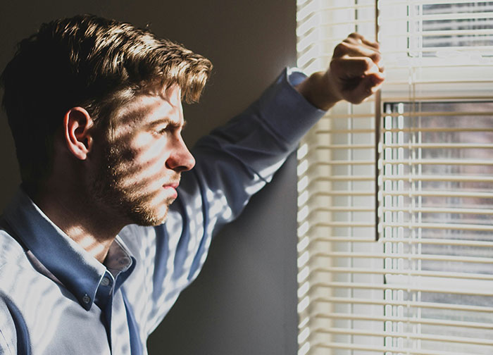 Young man looking out window blinds with a serious expression, reflecting on relationship conflicts about closing closet door.
